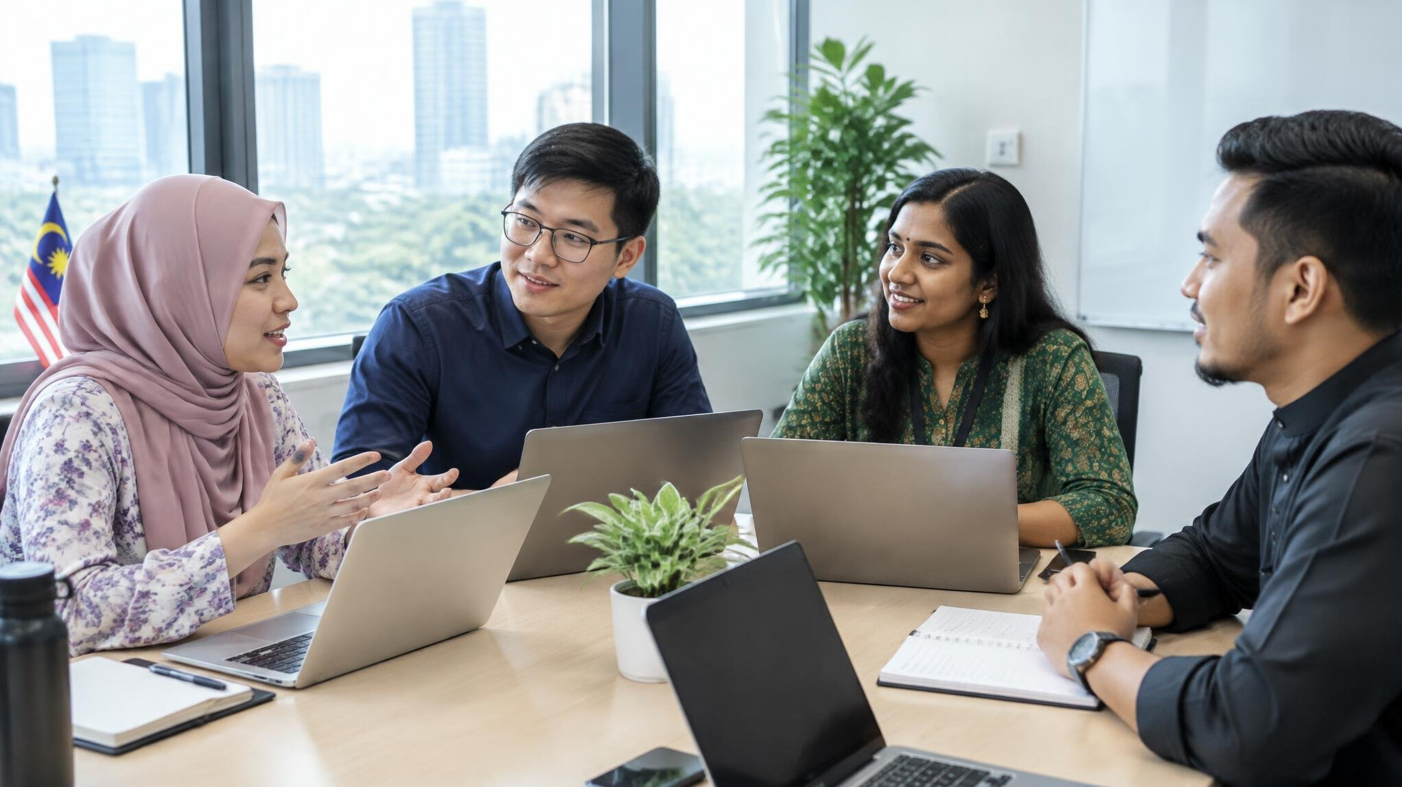 Diverse Malaysian colleagues in a meeting room discussing around a table with laptops